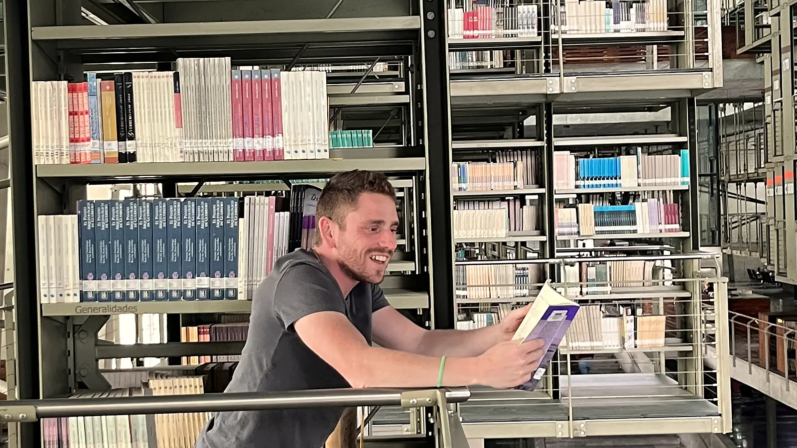 Yann Darmon reading, leaning on a rail in a library full of open stacks